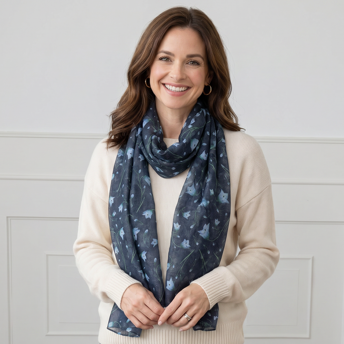 Woman wearing a blue floral patterned scarf, white sweater, and blue jeans against a white paneled wall.