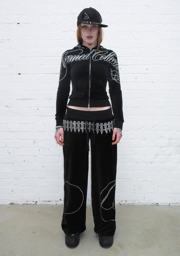 Model wearing a black velour hoodie with rhinestone embellishments and bullet design, paired with matching pants.