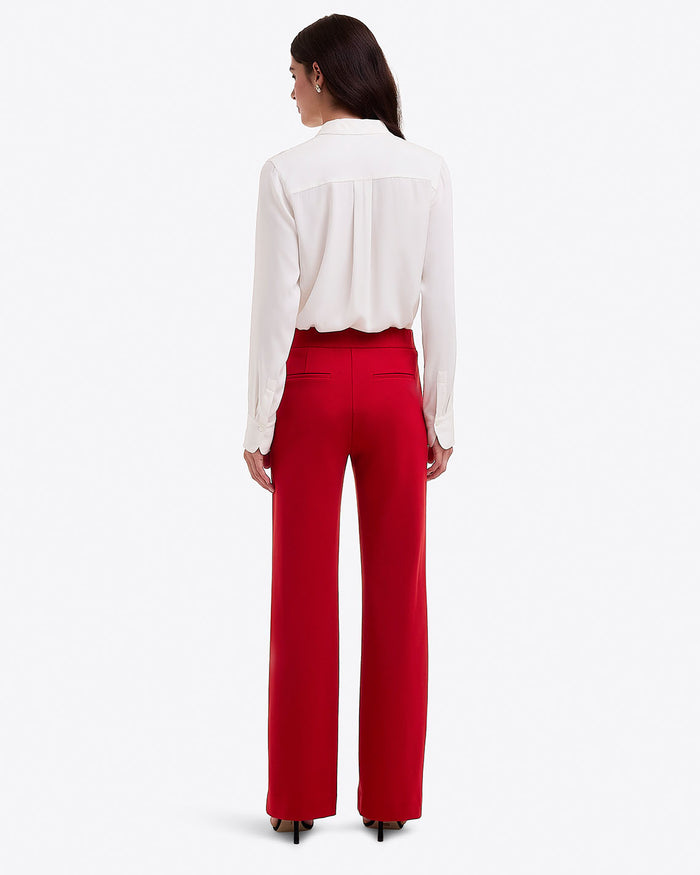 A woman faces away against a white background, wearing Draper James Flare Pants in Ponte in a lipstick shade, paired with a long-sleeve white blouse and black heels.