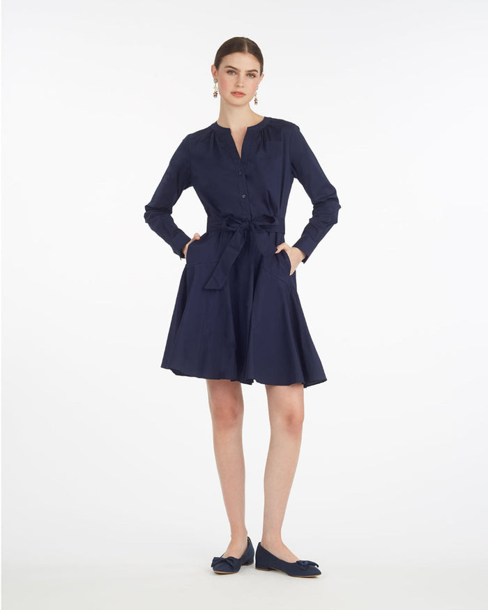 A woman stands against a white background wearing the Draper James Imogen Mini Shirtdressâ€”an effortless navy blue cotton work dress with long sleeves, a waist tie, and matching flats.