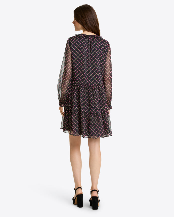 A woman faces away, wearing the Draper James Lorrie Long-Sleeve Dress in Crinkle Chiffonâ€”a knee-length black dress with a geometric pattern and ruffled hemâ€”paired with black high-heeled sandals against a plain background.