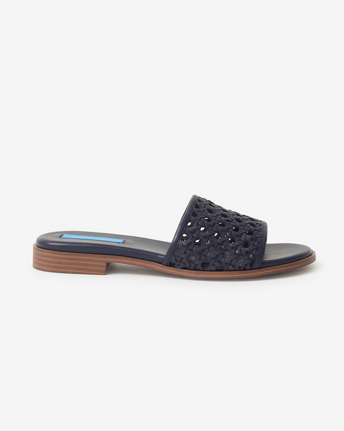 The Draper James Rattan Sandal is shown against a white background, featuring a woven navy blue upper, flat sole, and low wooden heel.