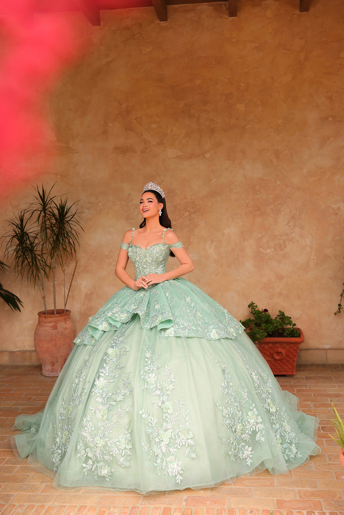 A young woman in an avocado green, embroidered ball gown with a tiara stands confidently on a brick floor.
