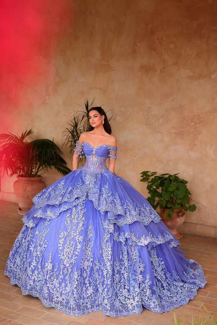 A woman wears a dramatic periwinkle/light blue tiered ball gown with intricate lace and off-shoulder design.
