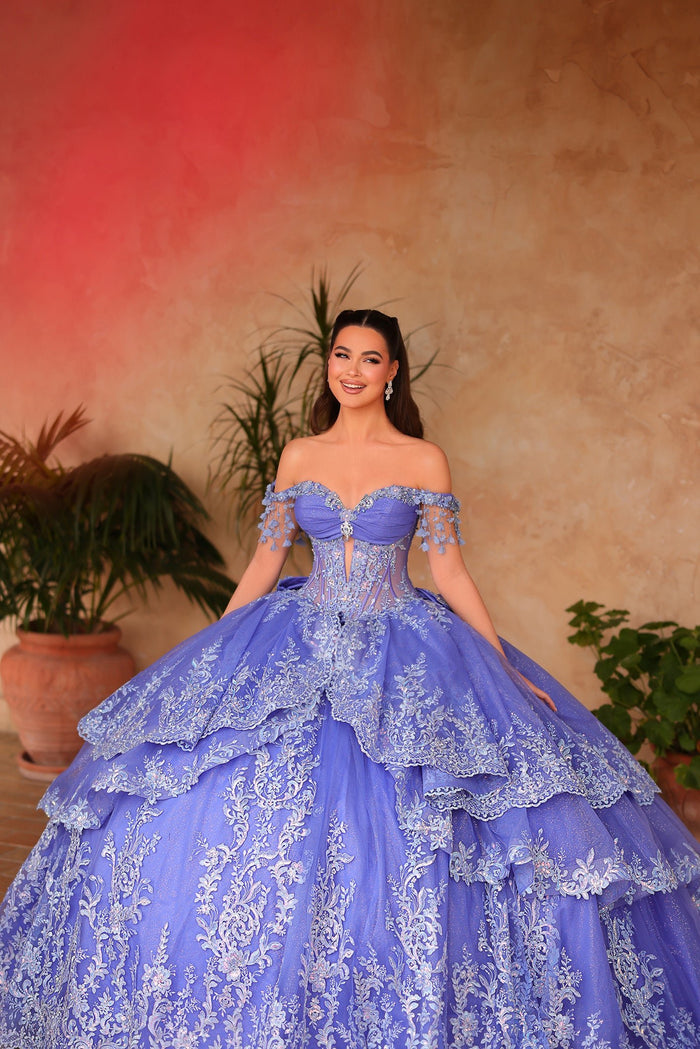 A woman in a lavish periwinkle blue quinceañera gown with intricate lace and off-shoulder design poses elegantly.