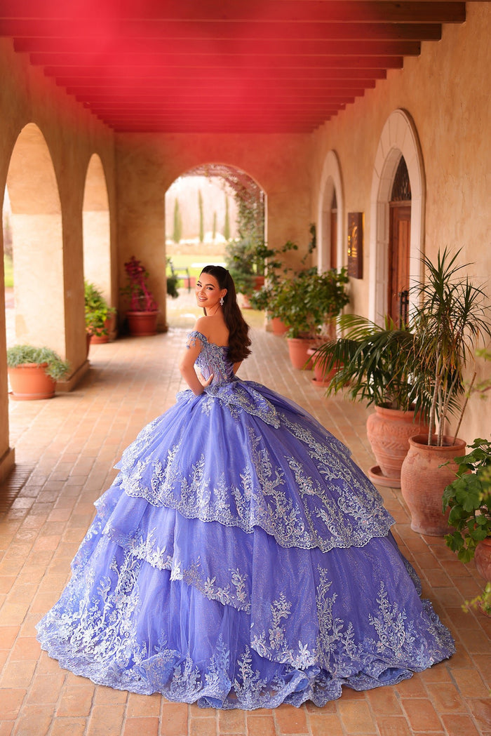 A woman in a lavish periwinkle quinceañera gown poses in an arched terracotta courtyard with plants.