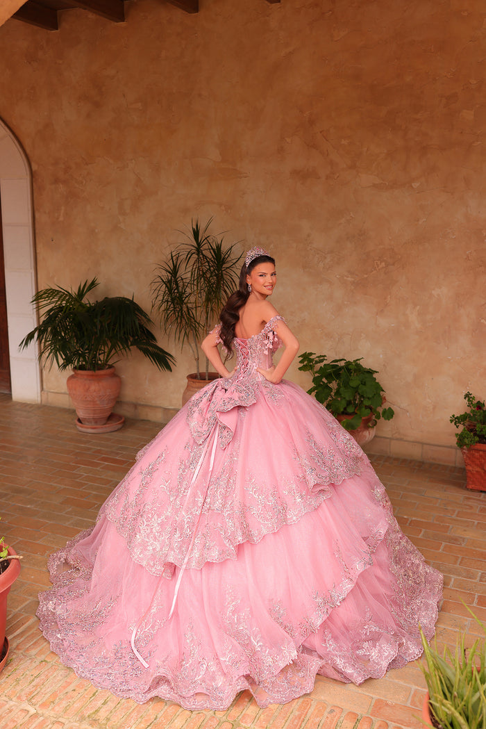 A young woman in a pink, silver-detailed ball gown poses in a terracotta-walled courtyard with potted plants.