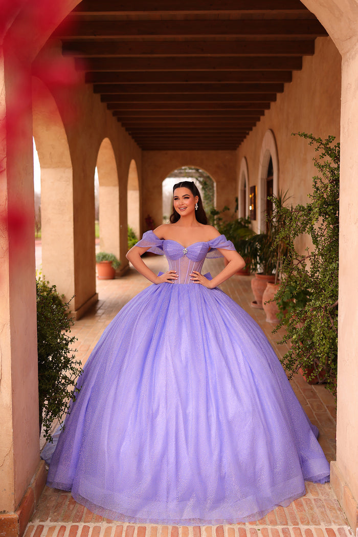 A woman wears a lilac off-shoulder ball gown in an arched stone hallway with potted plants.