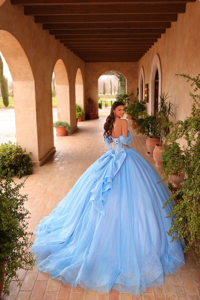 A woman in a voluminous blue ball gown stands in an arched terracotta corridor with potted plants.