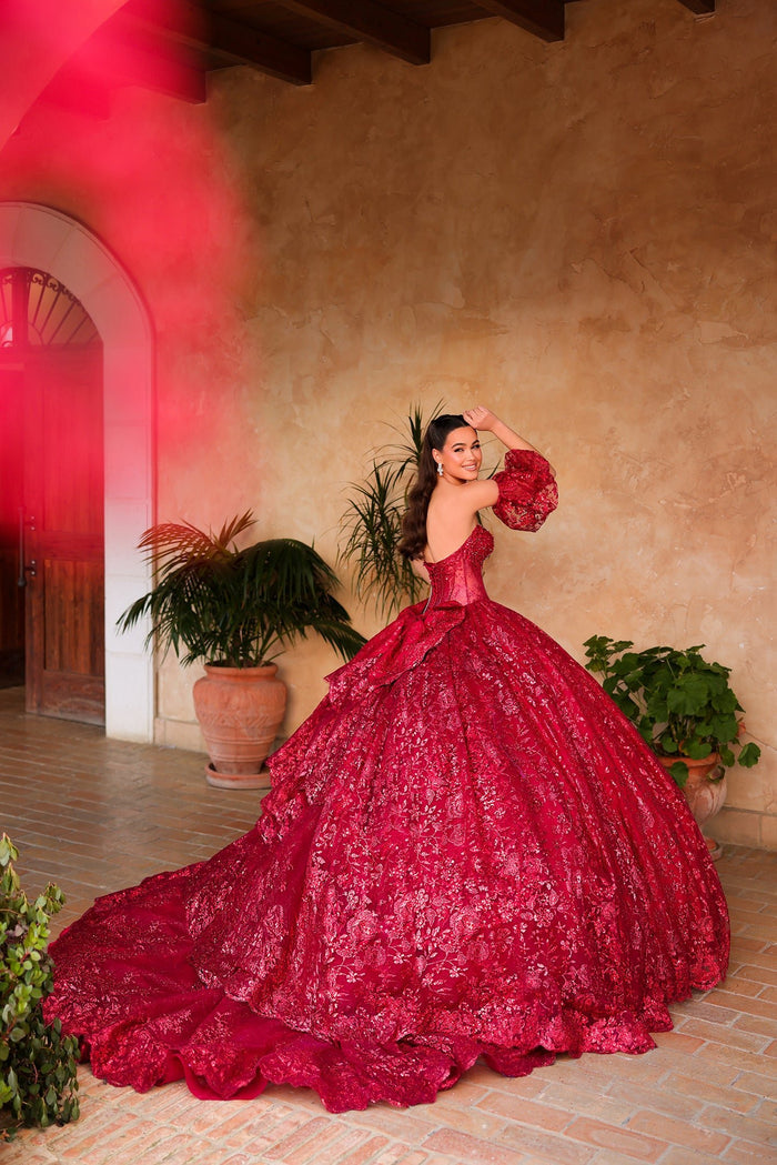 A woman in a vibrant red ball gown poses dramatically in a rustic terracotta-walled courtyard with potted plants.
