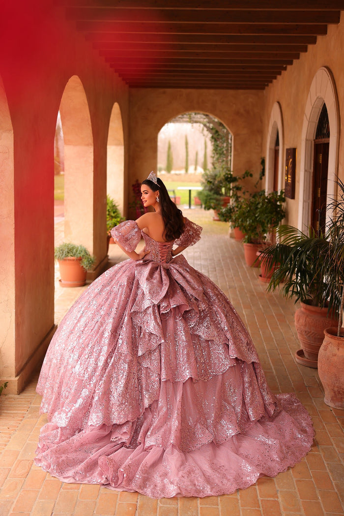 A woman in a sparkling pink ball gown with a tiara stands in an arched Mediterranean-style hallway.