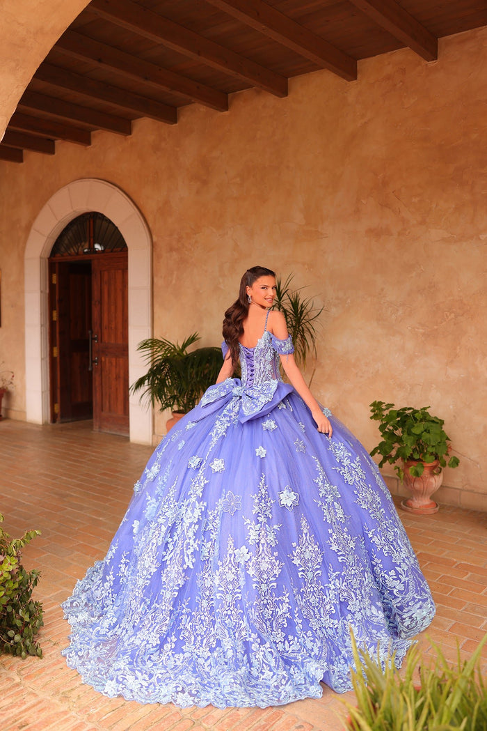 A woman in a lavish periwinkle blue ballgown with floral lace stands in a rustic Spanish-style courtyard.