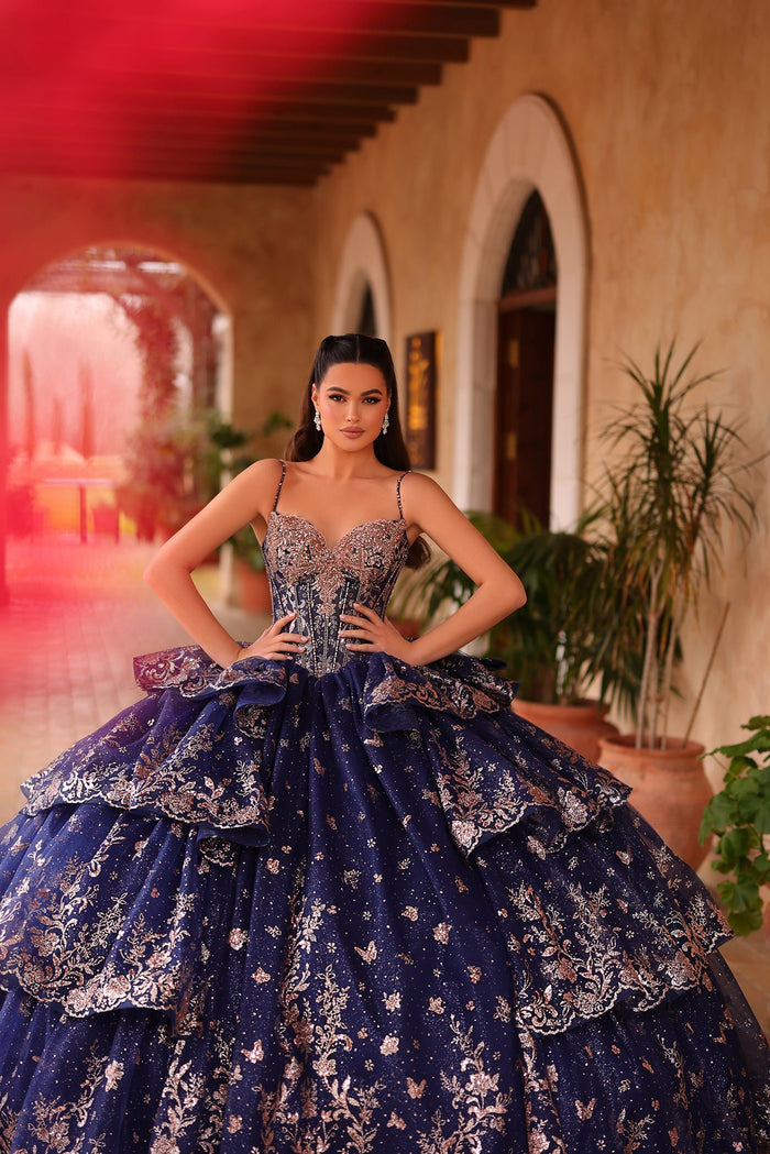 A woman in a navy blue, intricately embroidered ball gown with butterfly motifs poses in an arched hallway.