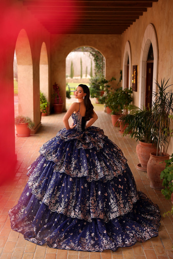 A woman in a navy blue and silver quinceañera gown stands in a sunlit Mediterranean-style courtyard.