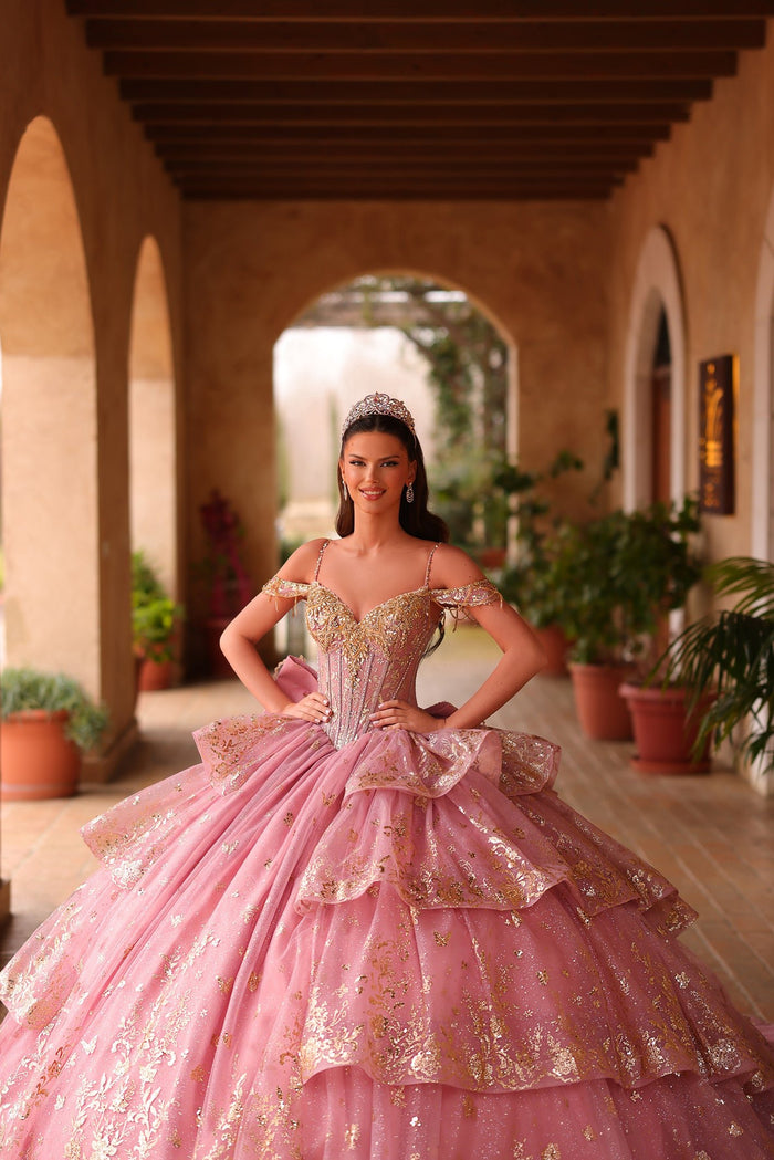 A young woman wears a pink and gold sparkly quinceañera ball gown with a tiara in an arched courtyard.