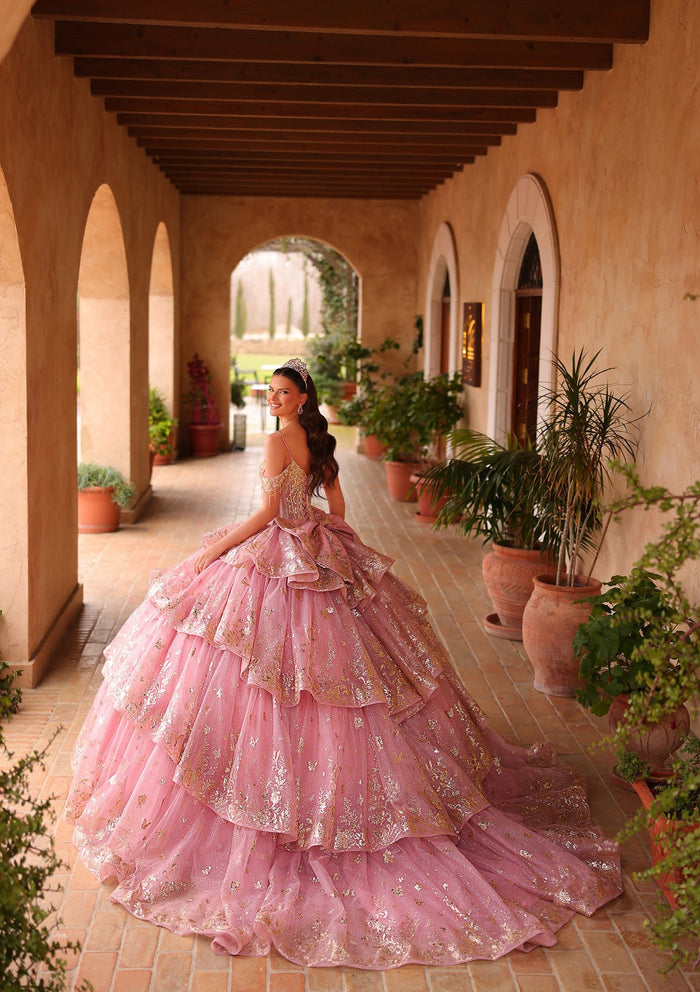A woman in a voluminous pink quinceañera gown poses in an arched Mediterranean-style courtyard with potted plants.