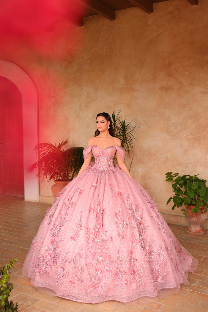 A woman wears an elaborate dusty rose off-shoulder ball gown with floral embroidery in a terracotta-toned room.