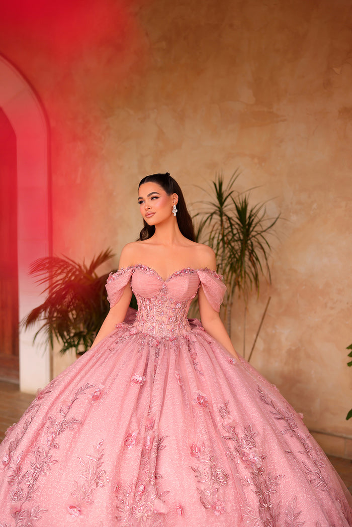 A woman wears an ornate pink off-shoulder ball gown with floral embroidery against a warm-toned background.