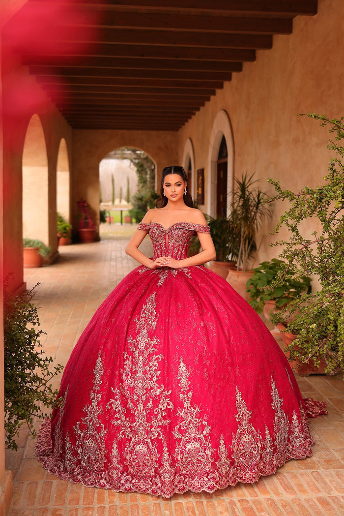 A woman wears an elaborate wine-colored quinceañera ball gown with intricate silver embroidery in a sunlit courtyard.