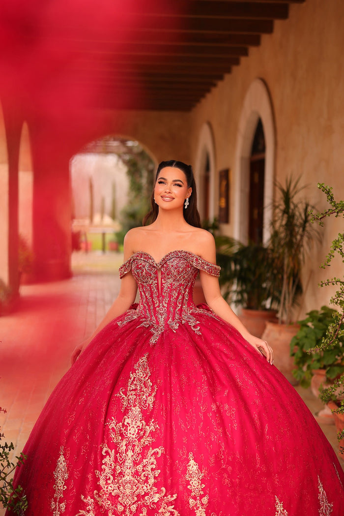 A woman wears an ornate red ball gown with silver embroidery, standing in an arched hallway with plants.