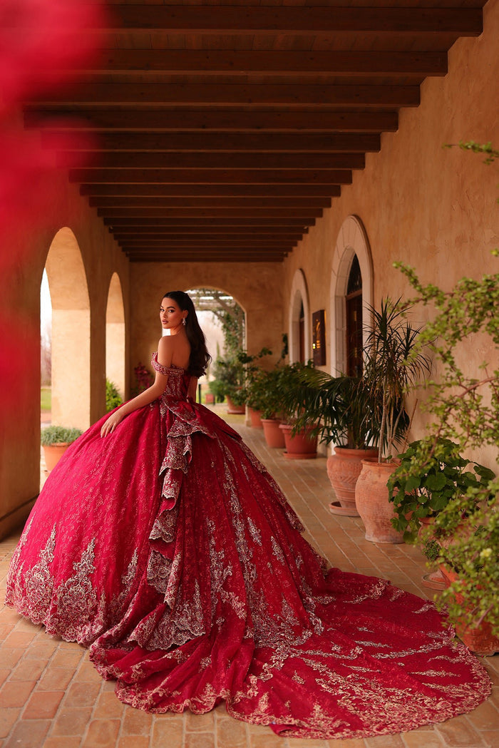 A woman in a vibrant red quinceañera gown stands in an arched Mediterranean-style corridor with potted plants.