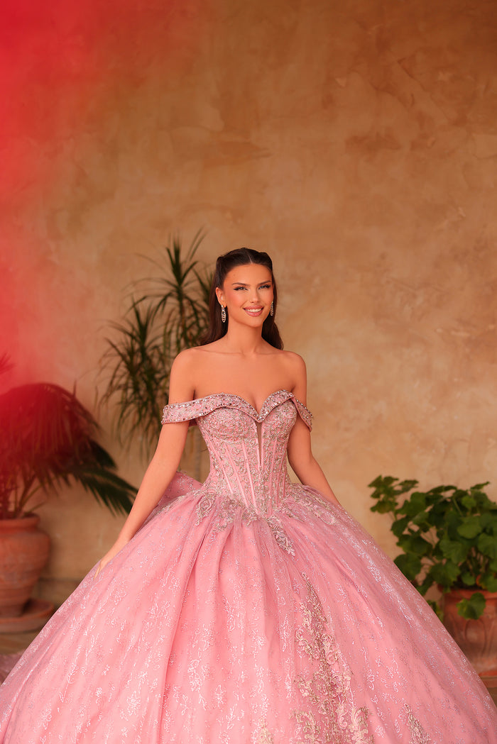 A woman wears a pink sparkly off-shoulder ball gown against a warm-toned background with potted plants.