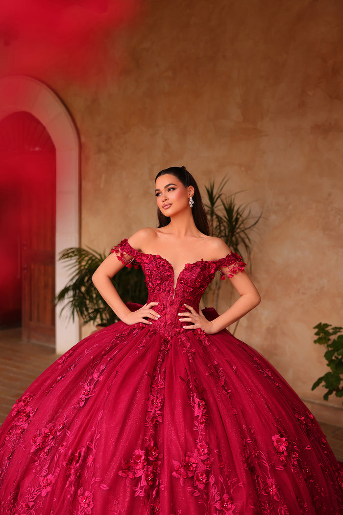 A woman wears a vibrant red off-shoulder ball gown with 3D floral appliqués in an arched, warm-toned setting.