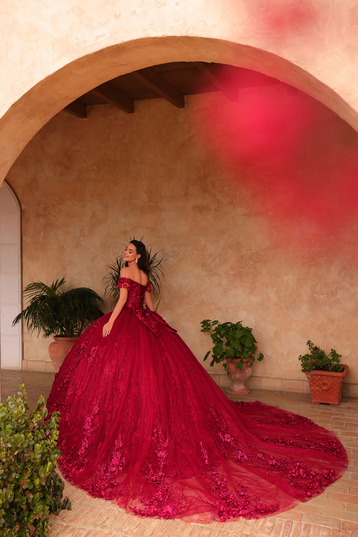 A woman in a voluminous red ball gown poses under an archway with potted plants and terracotta tiles.