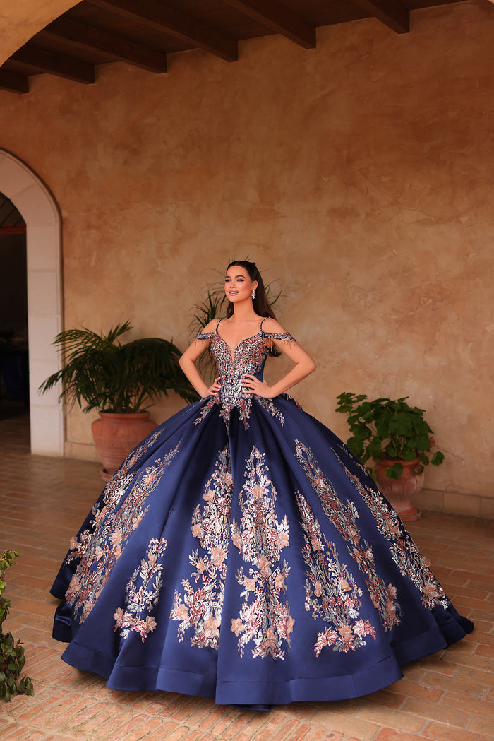 A woman in a navy ball gown with floral embellishments poses confidently in a warm-toned, arched hallway.