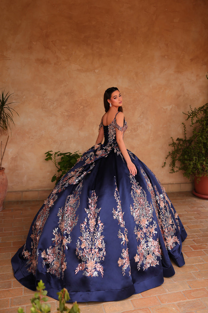 A woman in a voluminous navy blue ballgown with floral embroidery stands against a warm terracotta wall.