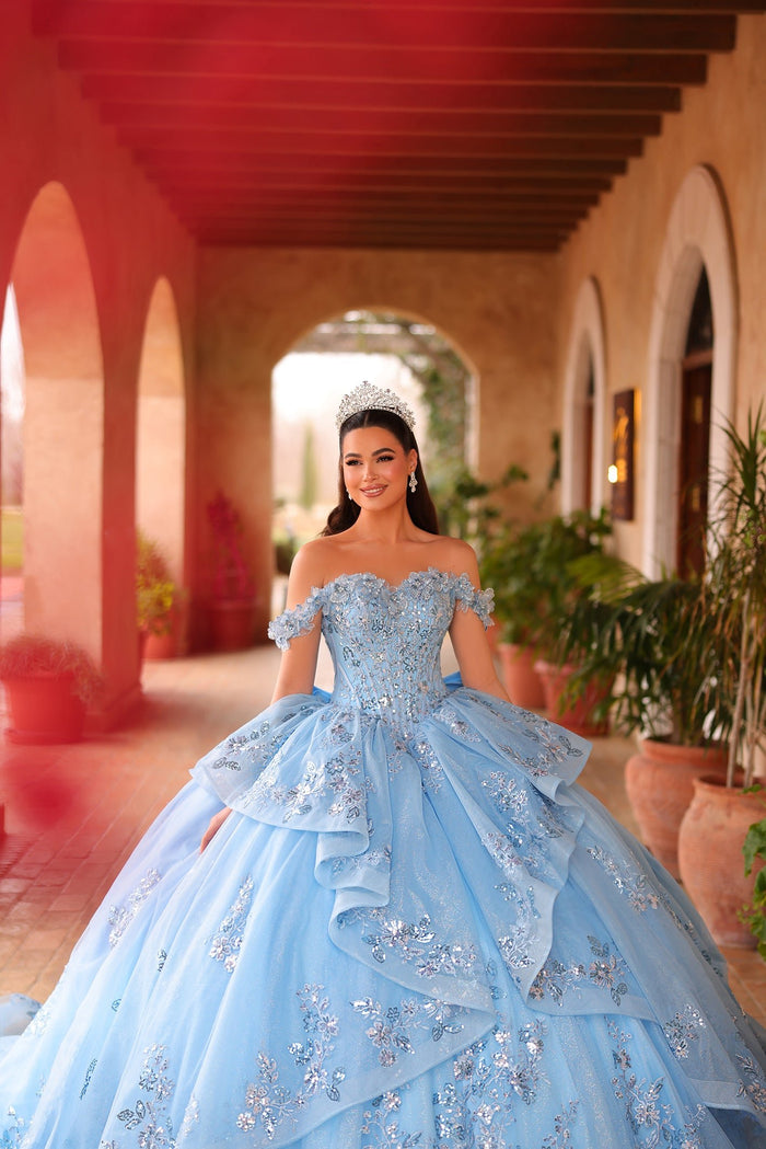 A young woman in a blue floral ball gown and tiara stands in an arched hallway with terracotta pots.