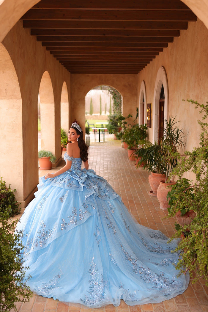 A woman in a blue, sparkly ball gown with a tiara poses in an arched Mediterranean-style hallway.