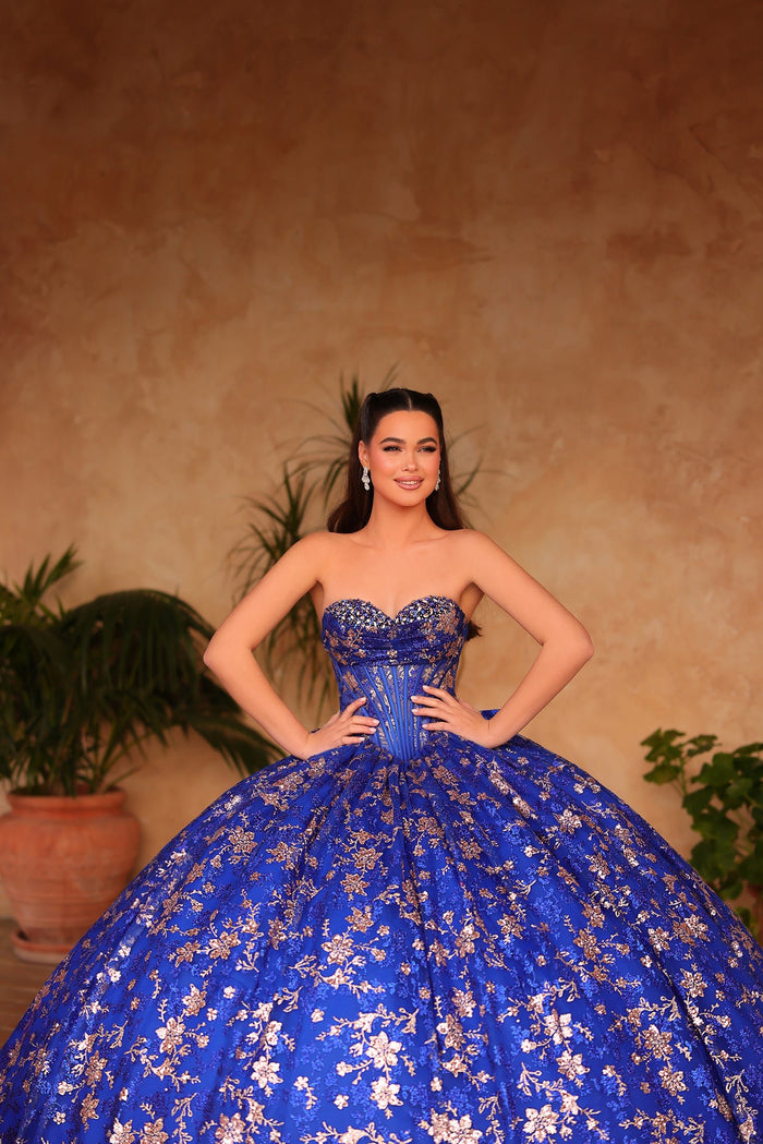 A woman wears an elaborate royal blue ball gown with floral embroidery, posing confidently against a warm background.