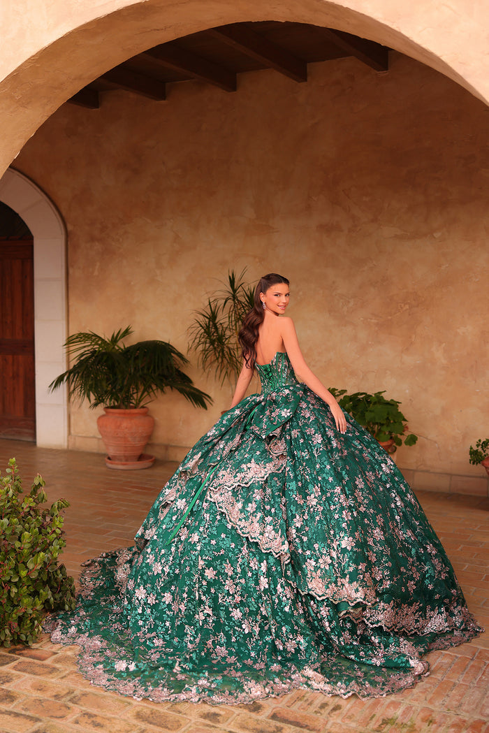 A woman in an emerald green floral ballgown poses dramatically in an arched, terracotta-walled courtyard.