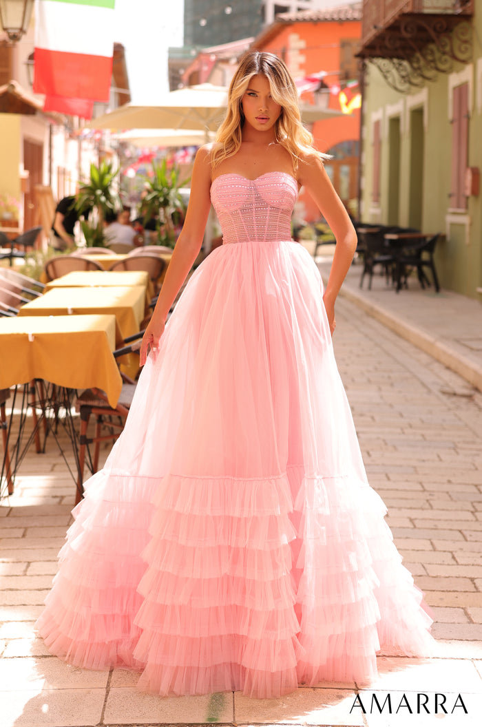 A woman wears a strapless, voluminous light pink tulle ball gown standing on a cobblestone street with cafe tables.