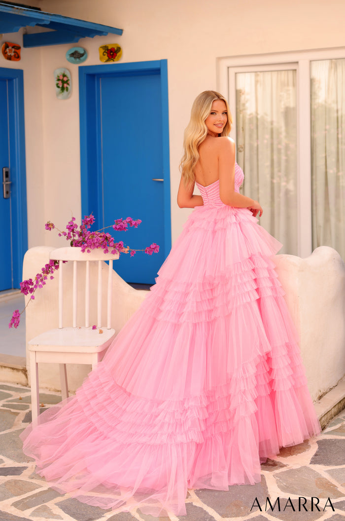 A blonde woman wears a voluminous pink tiered ball gown against a blue-doored Mediterranean-style backdrop.