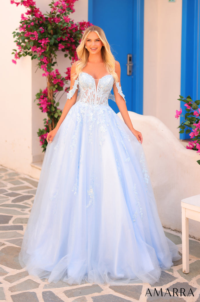 A model wears a light blue, off-shoulder ball gown with floral appliques against a blue door and pink bougainvillea.