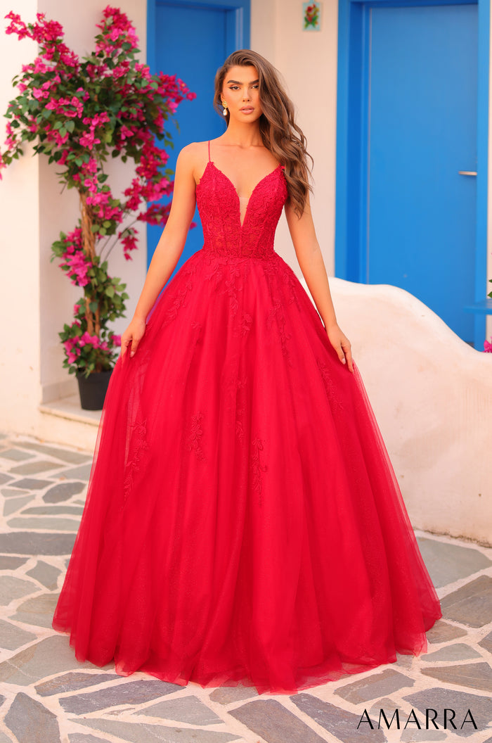 A model wears a full red ball gown with a fitted bodice and tulle skirt, standing against a blue door and pink flowers.