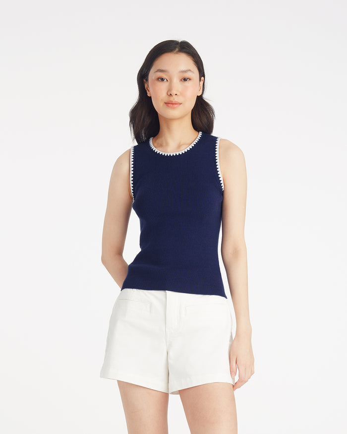A woman stands against a plain background wearing the Draper James Sweater Tank in navy with white trim, paired with white shorts.