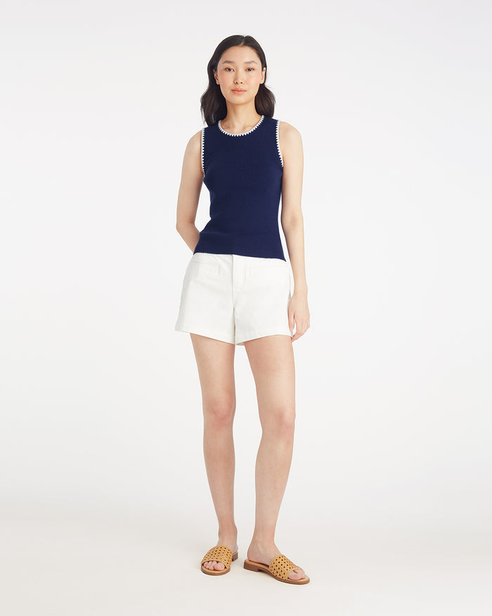 A woman stands against a plain white background wearing the Draper James Sweater Tank in navy with white trim, paired with white shorts and tan woven slide sandals.