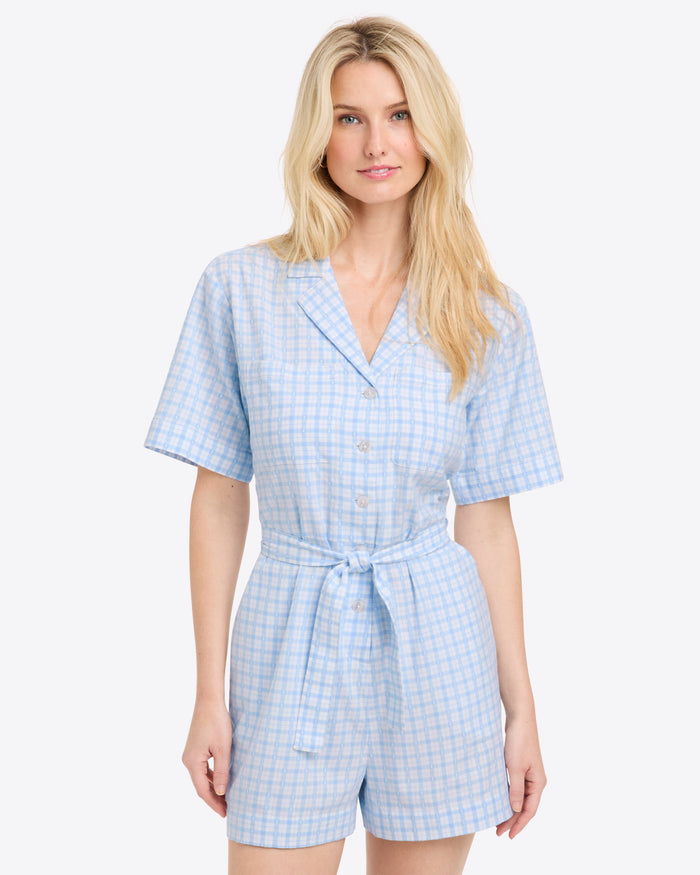 A woman wears the Draper James Tie Waist Romper, a light blue and white gingham one-piece with short sleeves, button front, and tied waist, standing against a plain light background.