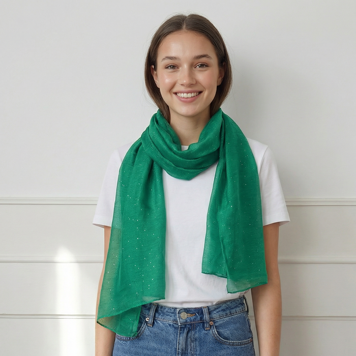 Woman wearing a green scarf, white shirt, and blue jeans against a white wall.